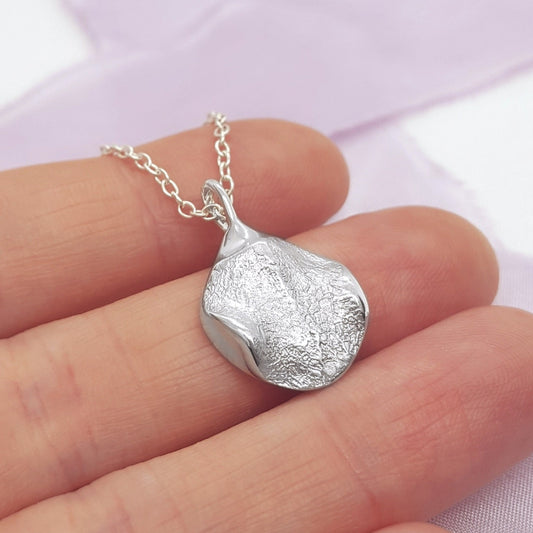 A silver pendant necklace with a textured, organic teardrop shape, held on a person's fingers against a soft lavender fabric background.