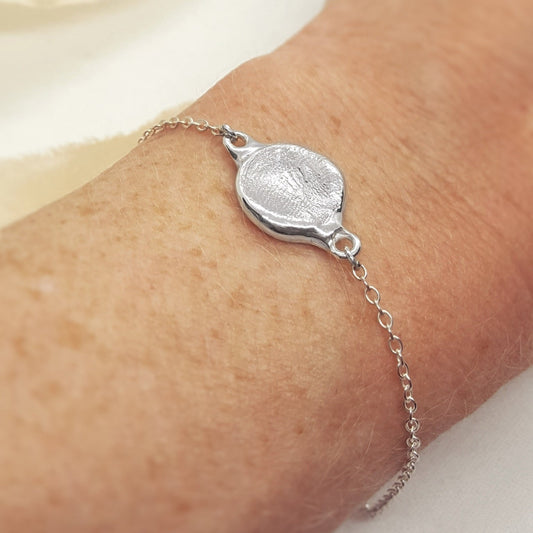 A close-up of a delicate silver chain bracelet with a textured oval medallion charm, worn on a person's wrist.