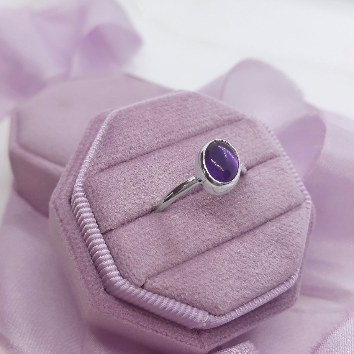 Silver ring with a purple gemstone in a matching purple box on a soft pink background