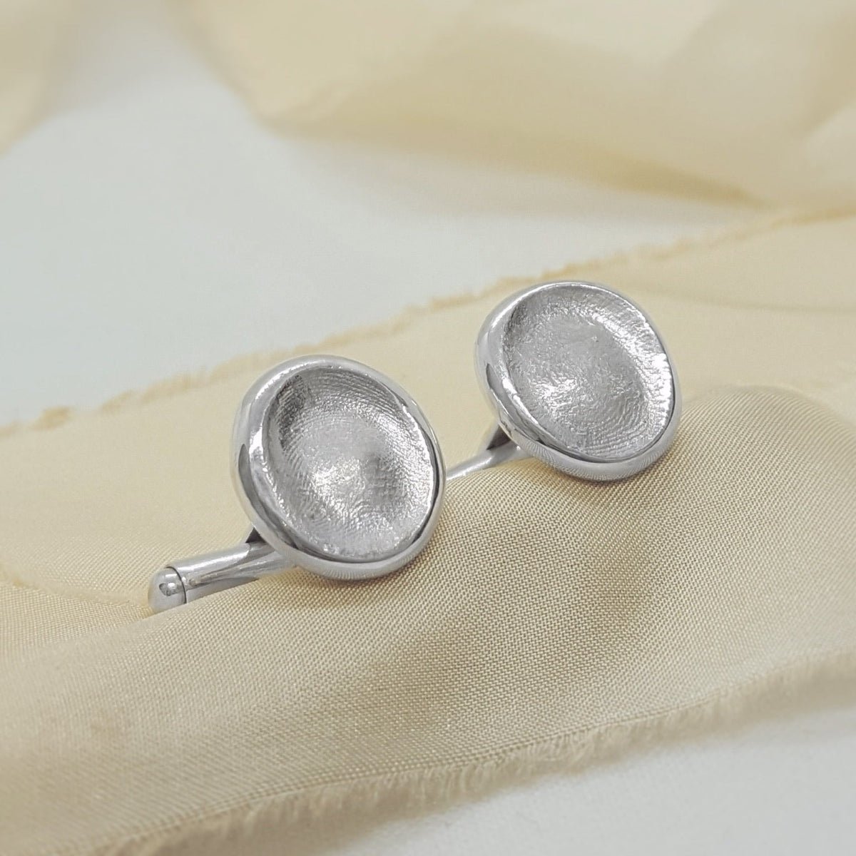 A pair of silver oval cufflinks with a textured, hammered finish, displayed on a soft beige fabric background.