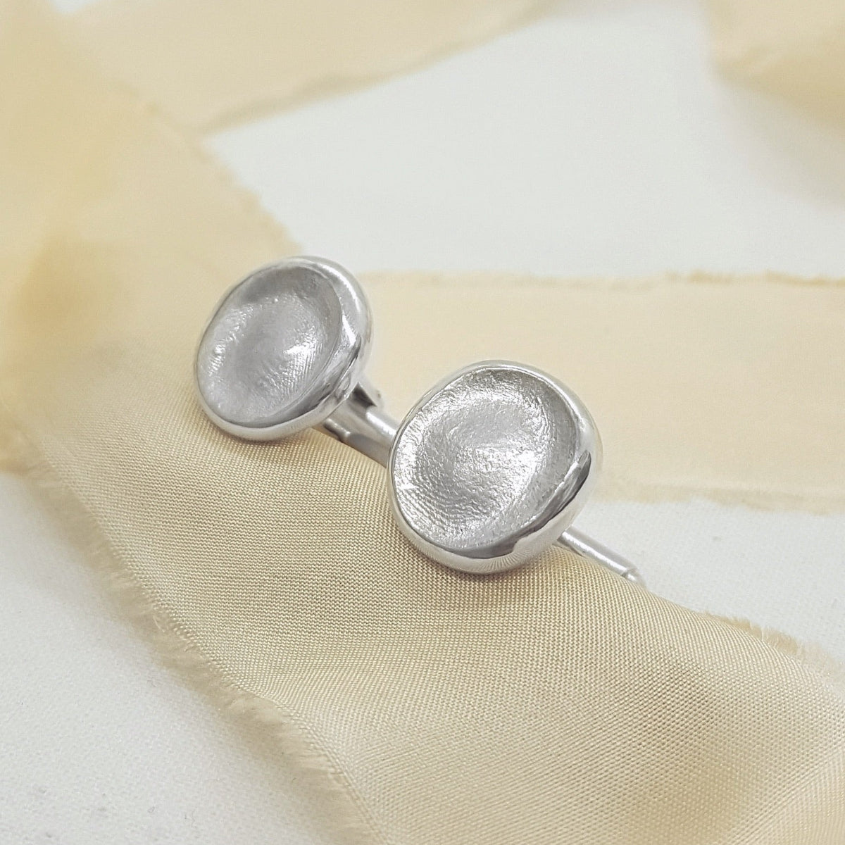 A pair of textured silver cufflinks displayed on a soft beige silk ribbon against a light cream background.