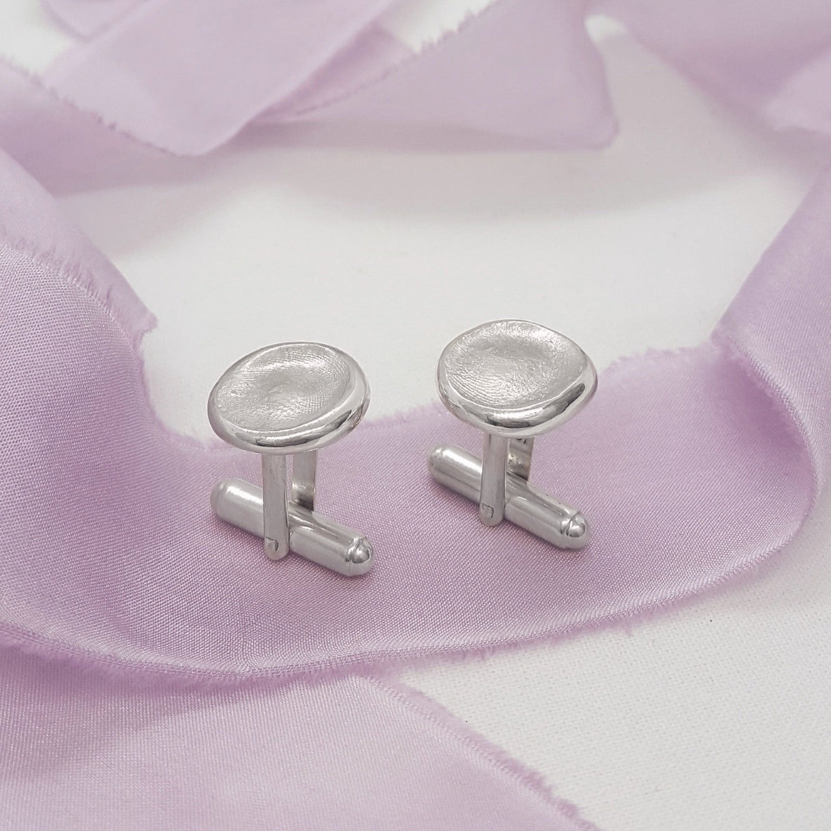 A pair of silver-toned metal cufflinks with oval faces, displayed on a soft lavender ribbon over a white background.
