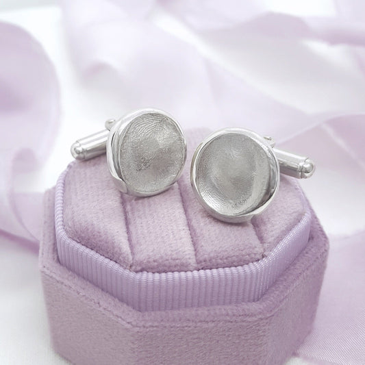 A pair of silver-toned oval cufflinks displayed on a lavender ribbed jewelry box against a soft white and pale pink fabric background.