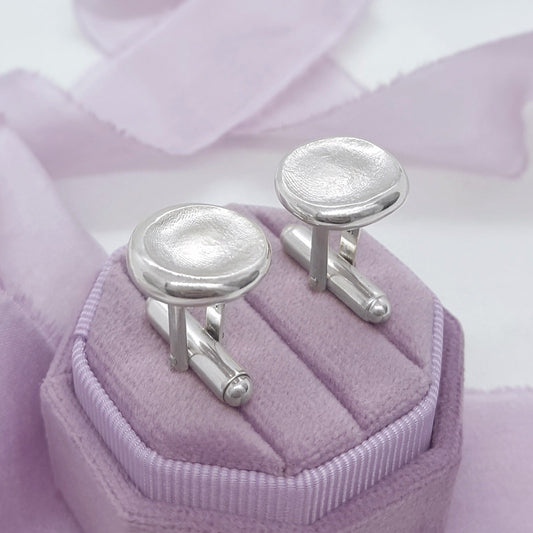 A pair of silver-toned oval cufflinks displayed on a lavender jewelry box, with soft lavender ribbon in the background.