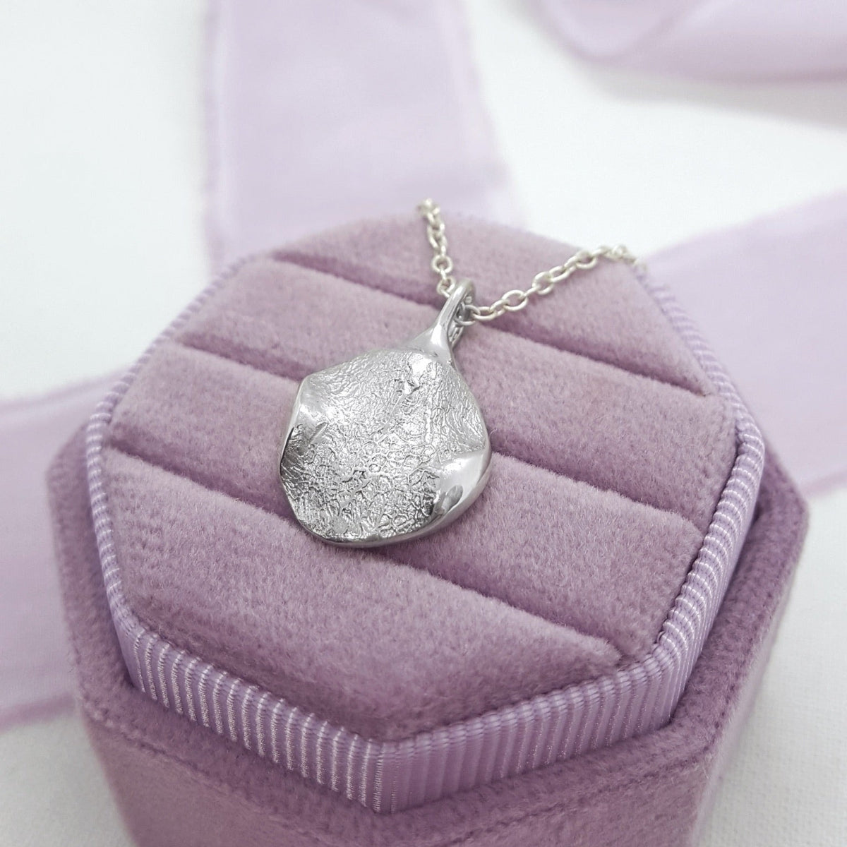 A silver textured pendant necklace resting on a lavender purple velvet jewelry box.
