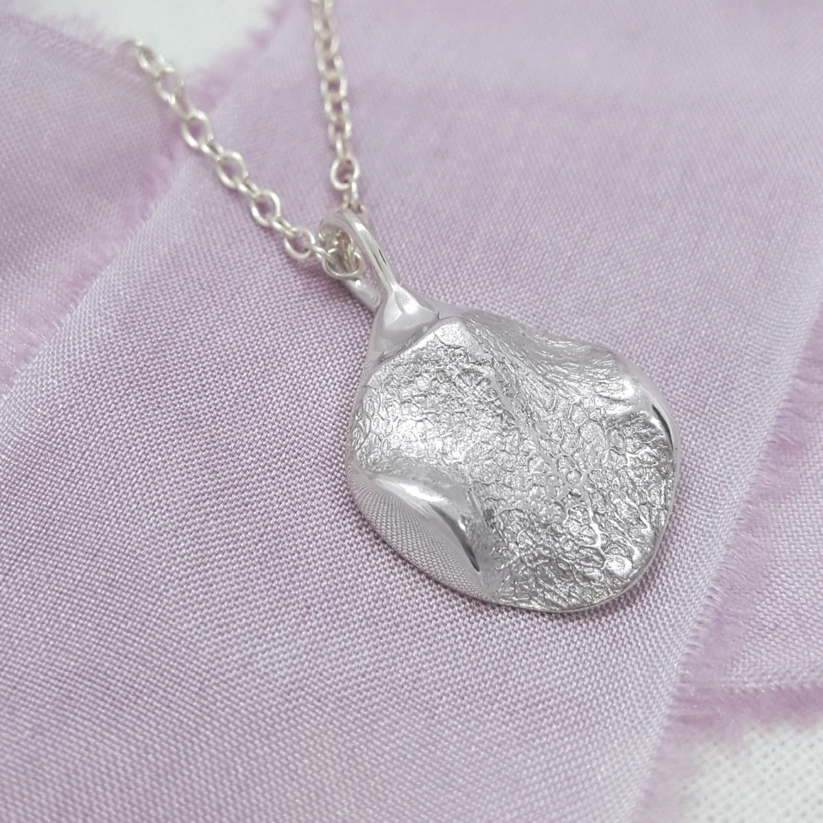 A textured sterling silver pendant necklace with an organic, irregular shape resting on a soft lavender fabric background.