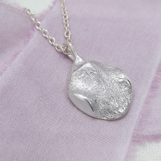 A textured sterling silver pendant necklace with an organic, irregular shape resting on a soft lavender fabric background.