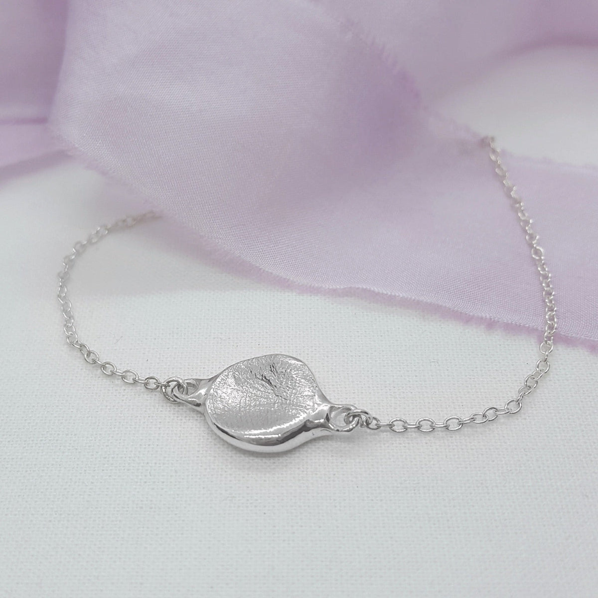 A delicate silver chain bracelet featuring a textured, organic-shaped silver pendant, displayed on a white surface with a soft lavender fabric backdrop.