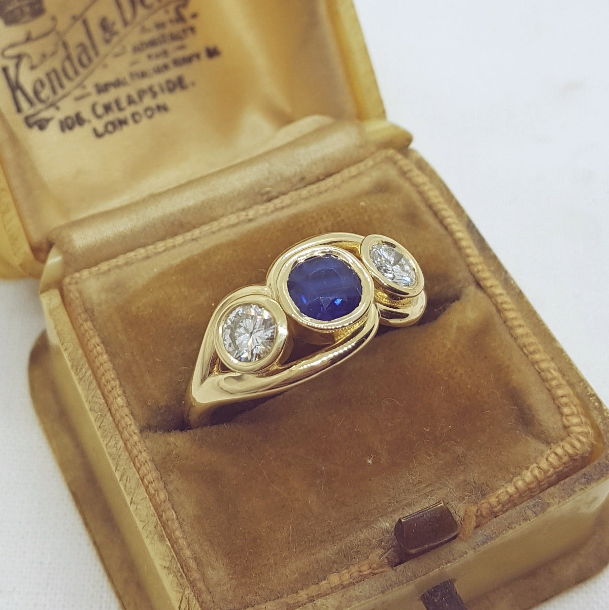 Gold ring with a blue gemstone and diamonds on a brown velvet cushion with 'Kendal & Douglas' branding.