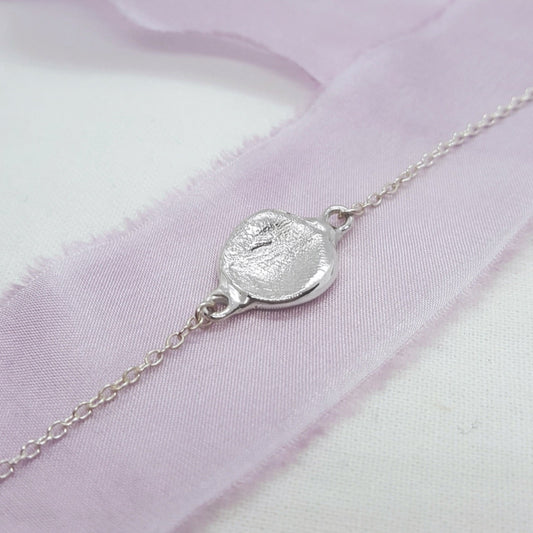 A delicate silver chain bracelet with a textured, irregularly shaped pendant, displayed on a soft pink fabric ribbon over a white background.