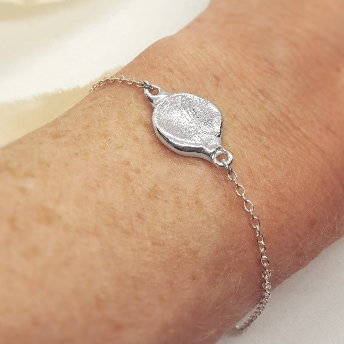 A close-up of a delicate silver chain bracelet with a textured oval medallion charm, worn on a person's wrist.