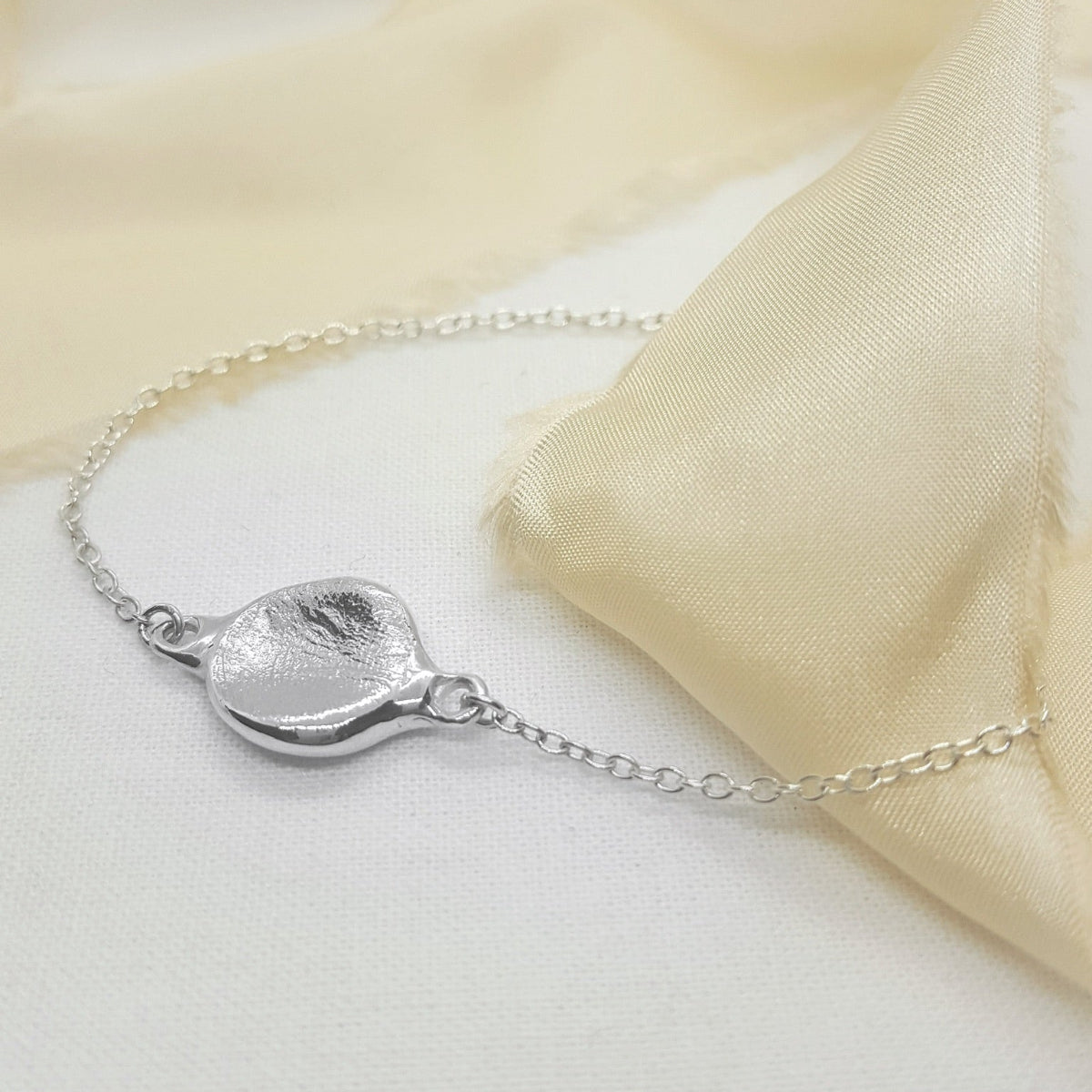 A silver chain bracelet with a textured, organic-shaped pendant, styled on a white surface with a soft cream-colored satin fabric drape.