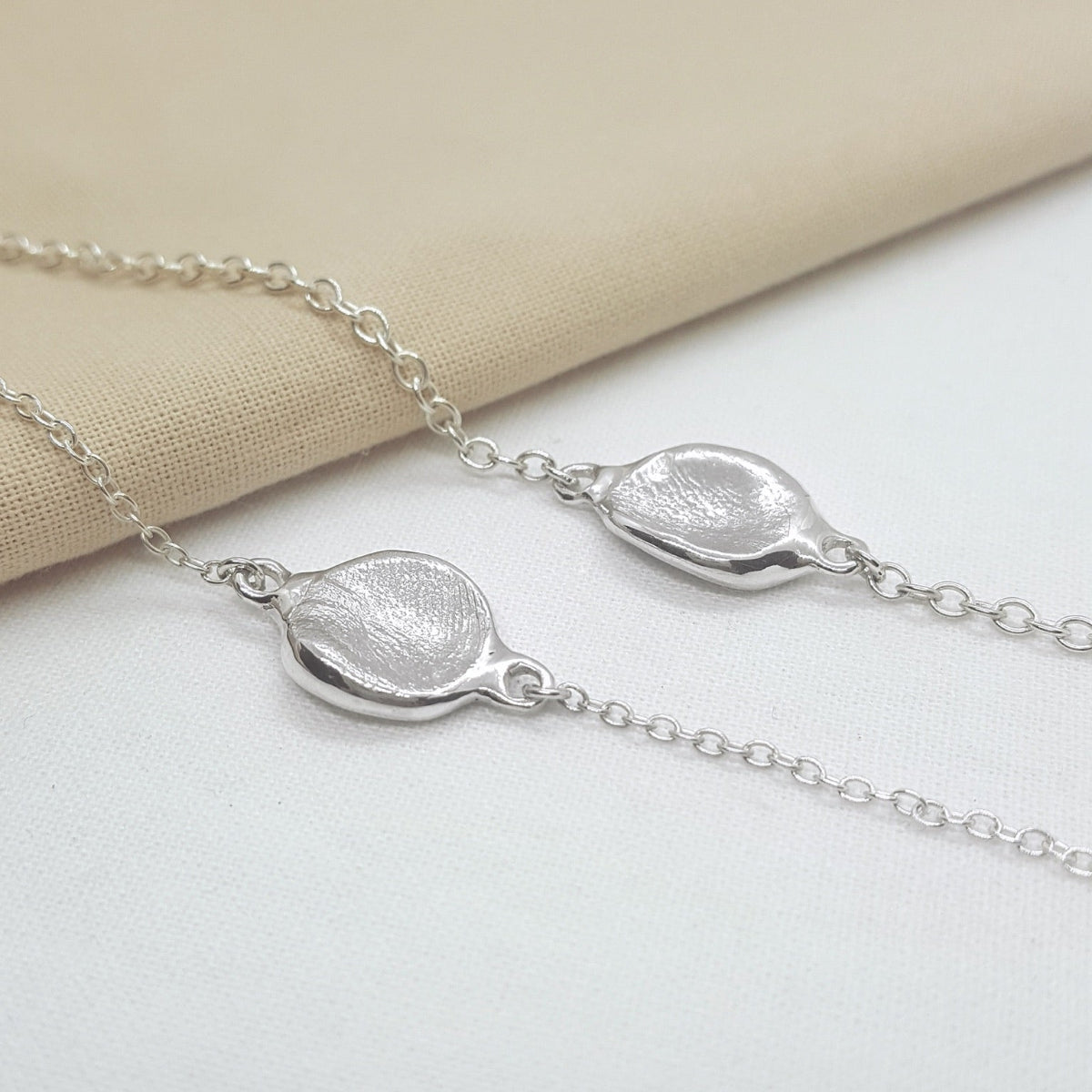 A close-up of a silver necklace with two textured, organic-shaped oval pendants on a delicate chain, resting on a beige fabric and white surface.