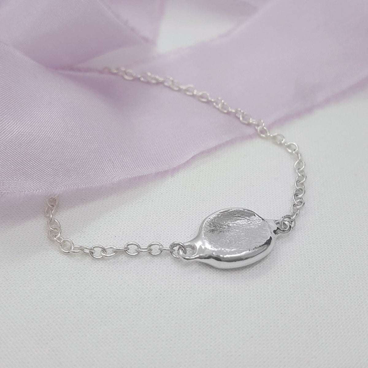 A silver chain bracelet with a textured, organic-shaped pendant, displayed on a white fabric background with a soft lavender ribbon.