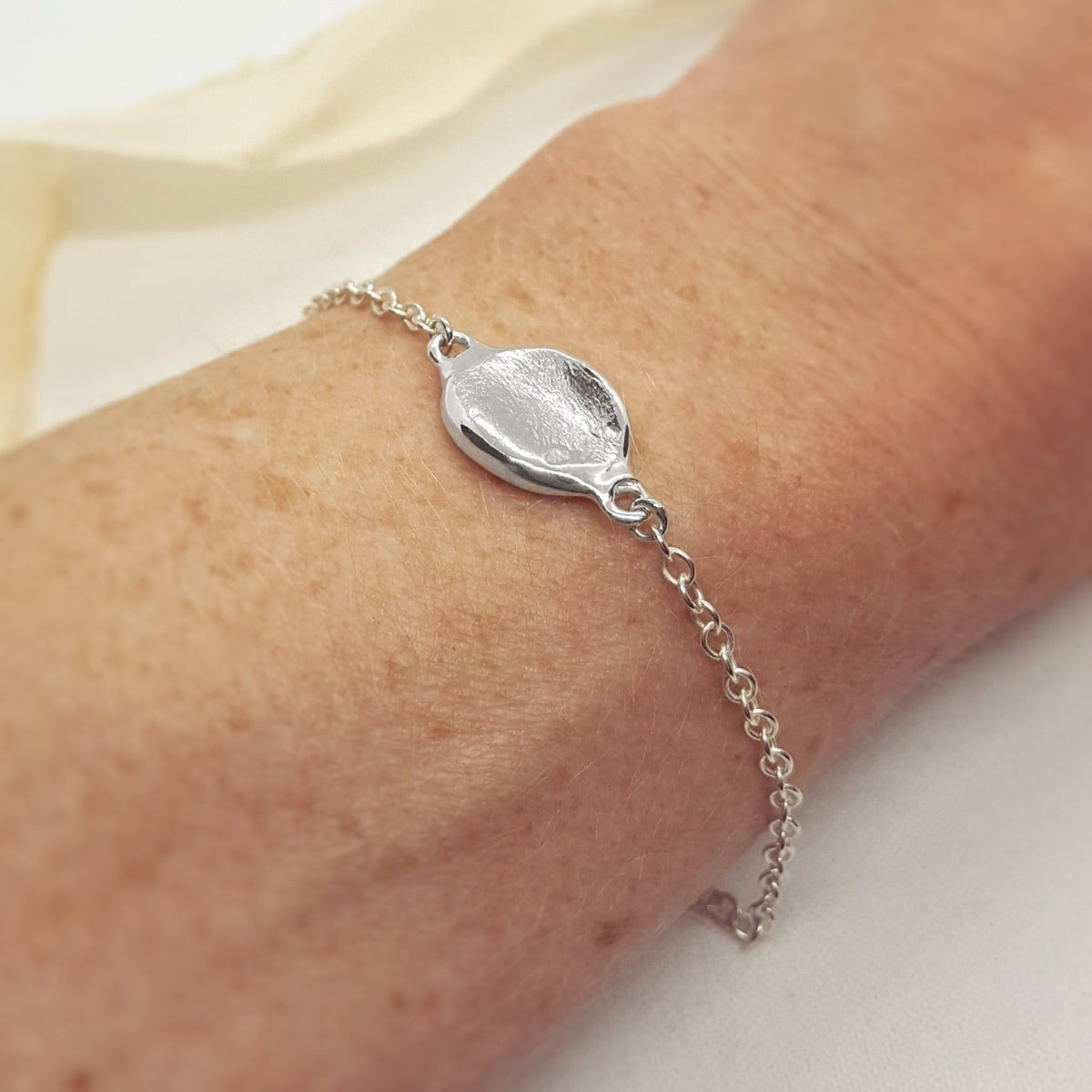 A close-up of a delicate silver chain bracelet with a textured, organic-shaped disc charm, worn on a person's wrist.